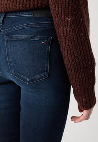 Dark blue denim jeans with a fitted design. Features include two rear pockets and a black waistband label. Brown, textured knitted sweater sleeve visible.