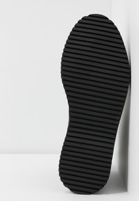 Suela de zapato negra con crestas horizontales espaciadas uniformemente, colocada verticalmente contra una esquina blanca de pared y suelo.