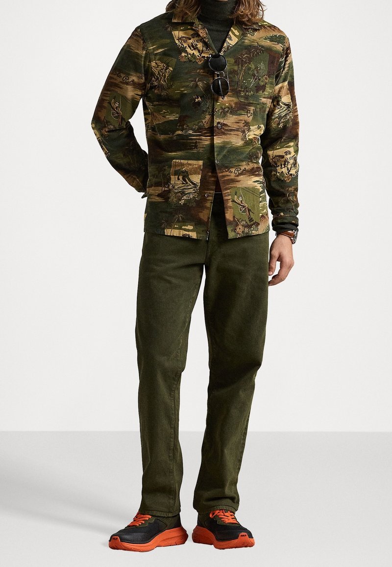 Homme portant un pantalon vert foncé, une chemise verte à motifs boutonnée avec un col roulé noir en dessous, et des chaussures noires avec des semelles et lacets orange vif.