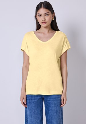 Gelbes kurzärmliges Oberteil mit V-Ausschnitt, aus einem leichten Stoff gefertigt. Kombiniert mit blauen weitem Jeans, steht das Model vor einem hellen Hintergrund.