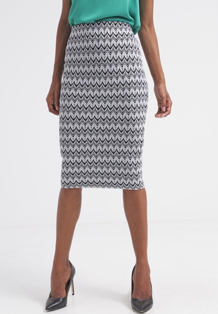 Femme portant une jupe crayon à motifs zigzag noirs et blancs longueur genou, un haut sans manches vert et des chaussures à talons noirs, debout.