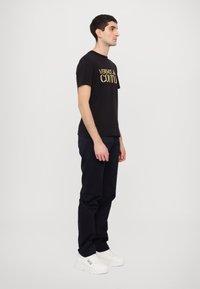 Juoda medvilninė marškinėliai su auksiniu užrašu "VERSACE JEAN COUTURE", derinami su tamsiai mėlynomis kelnėmis ir baltomis sportbačiais. Paprastas, modernus fasonas ir stilius.
