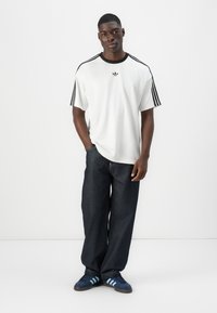 Witte t-shirt met korte mouwen en zwarte accenten, voorzien van drie zwarte strepen op de mouwen, gecombineerd met donkerdenim jeans en blauwgestreepte sneakers.