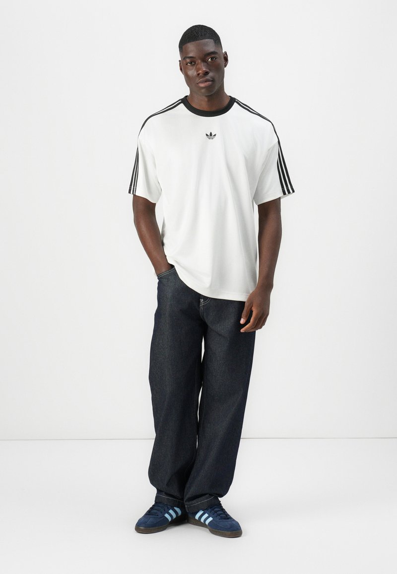 Witte t-shirt met korte mouwen en zwarte accenten, voorzien van drie zwarte strepen op de mouwen, gecombineerd met donkerdenim jeans en blauwgestreepte sneakers.