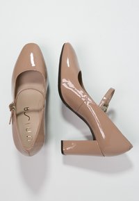 Beiga lackläder Mary Jane-skor med rund tå, ankelsnodd och fyrkantig klack. Slät textur med glänsande yta.