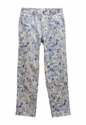 Frau steht mit den Händen in den Taschen, trägt eine blaue ärmellose Strickweste, gemusterte Jeans mit hoher Taille und blaue Mokassins.