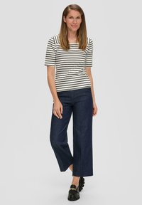 Gestreepte top met korte mouwen in wit en zwart, gecombineerd met een donkere denim wijde broek. Model draagt zwarte instapschoenen met gouden kettingdetails.