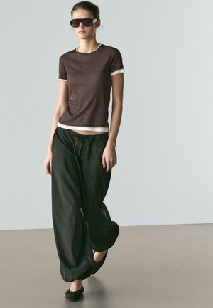 Woman walking wearing dark sunglasses, brown short-sleeve top over white shirt, loose black pants, and black flat shoes on gray floor.