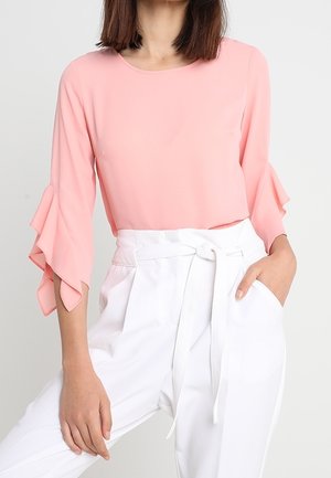 Vrouw met een lichtroze blouse met ruches aan de mouwen en een witte high-waist broek vastgeknoopt met een stoffen riem, hand in de zak.