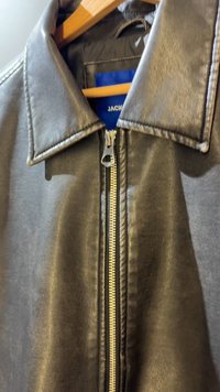 Chaqueta de cuero sintético negro con un cuello prominente y cierre frontal con cremallera y tirador plateado. Textura suave y líneas limpias.