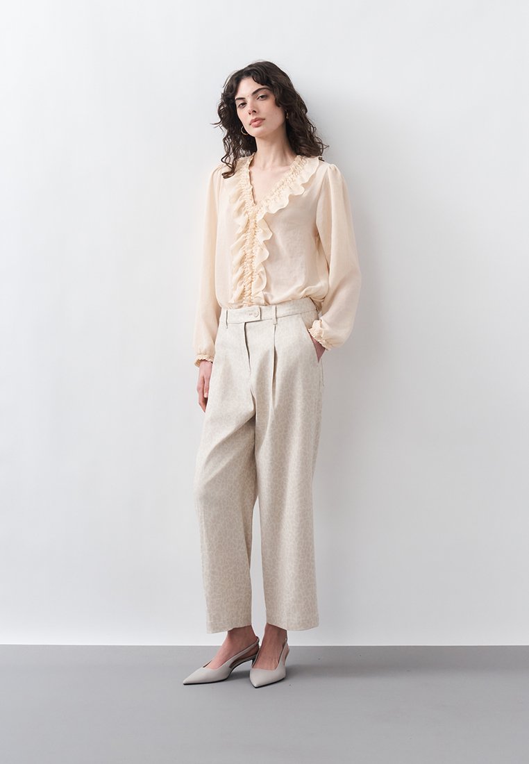 Femme debout contre un mur blanc, portant un chemisier crème à volants, un pantalon large taille haute et des escarpins pointus à bride arrière.