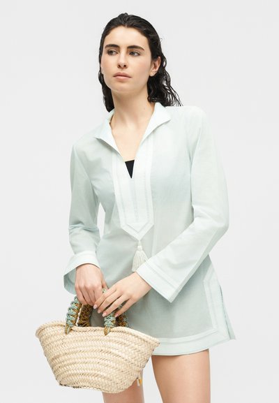 Γυναίκα με σκοτεινά μαλλιά που φοράει γαλάζια τουνίκ και κρατάει μια υφαντή τσάντα με πλεχτές λαβές, κοιτάζοντας προς το πλάι.