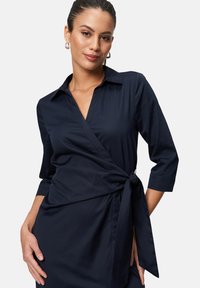 Navy blue wrap dress with a collar, three-quarter sleeves, and a tied waist detail. Smooth fabric, sleek design with minimal accents.