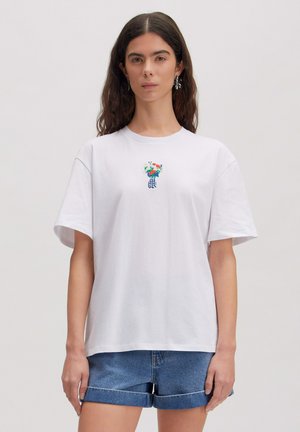 Woman with dark hair wears white T-shirt with small colorful flower bouquet design and blue rolled-up denim shorts.