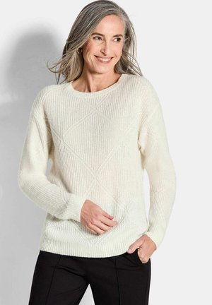 Frau mit grauen Haaren lächelt und trägt einen strukturierten weißen Strickpullover und schwarze Hose, posiert mit einer Hand in der Tasche vor einem schlichten Hintergrund.