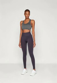 Egy nő szürke sportmelltartóban, magas derekú fekete leggingsben és fehér sportcipőben áll egy sima világos háttér előtt.