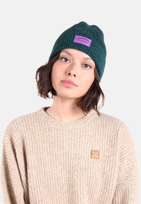 Bonnet en tricot vert foncé avec un revers et un patch logo violet ; porté avec un pull côtelé beige présentant une petite accentuation carrée.