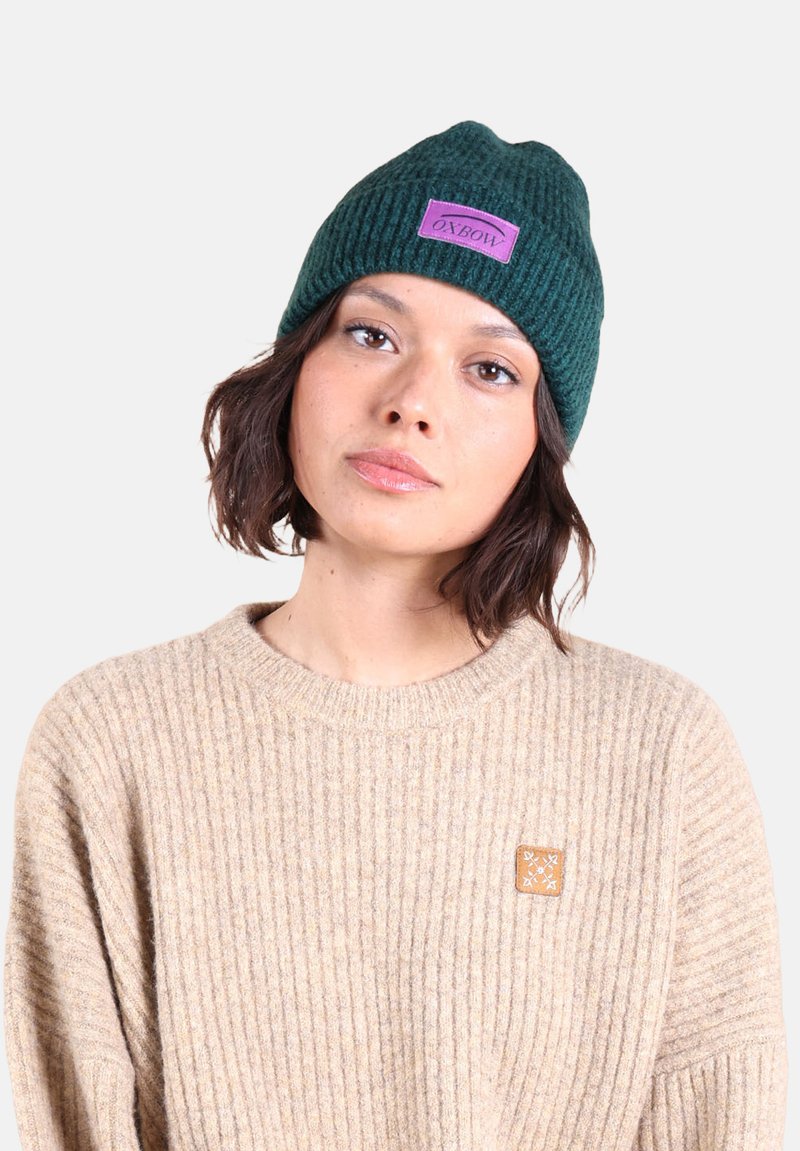 Bonnet en tricot vert foncé avec un revers et un patch logo violet ; porté avec un pull côtelé beige présentant une petite accentuation carrée.
