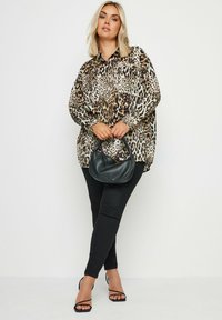 Bluzka w panterkę, luźny krój, satynowy materiał. Czarne dopasowane dżinsy, sandały na paski, czarna torebka. Jasne tło. Długie włosy.