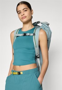 Türkiser Rucksack mit grauen Akzenten und verstellbaren Trägern, kombiniert mit einem passenden türkisen Oberteil. Glatte Textur, figurbetontes Design und ein gelbes Gürtel-Detail.