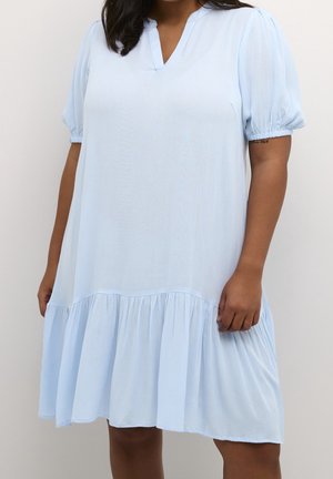 Femme portant une robe ample bleu clair avec des manches courtes bouffantes et un ourlet à volants, debout devant un fond neutre.