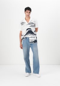 Kortermet hvit skjorte med svart fjell- og skyprint, kombinert med lyseblå vide jeans og hvite joggesko. Enkelt bakgrunn.