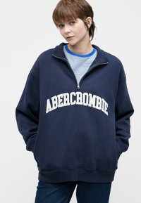 Granatowy sweter z białym napisem "ABERCROMBIE". Posiada kołnierz na zamek pół-zamknięty, luźny krój oraz ściągacze na mankietach. Noszony na niebieskiej, w paski koszuli.
