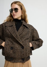 Dubbelzijdig herringbone jasje in bruin en zwart, met oversized revers, twee grote zwarte knopen en zakken aan de zijkanten.