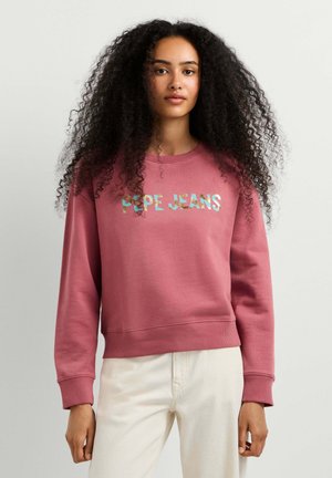Jeune femme aux longs cheveux bouclés portant un sweat-shirt rose avec un texte multicolore "PEPE JEANS" et un pantalon beige clair.