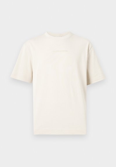 Κοντομάνικο, εκρού T-shirt με στρογγυλή λαιμόκοψη και διακριτικό ανάγλυφο λογότυπο στο στήθος, εμφανιζόμενο σε ανοιχτό γκρι φόντο.