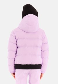 Persoon in een lichtpaarse donsjack en bijpassende broek met een zwarte beanie, van achteren gezien tegen een witte achtergrond.