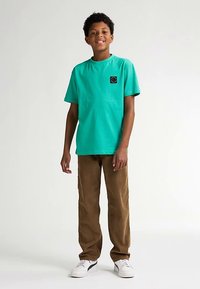 Turkoosi puuvillainen t-paita, jossa musta neliönmuotoinen logo, yhdistettynä ruskeisiin puuvillahousuihin ja valkoisiin lenkkareihin, malli seisoo rentoine käsivarsineen.