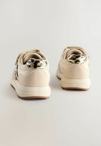 Une paire de baskets beiges avec des accents de motif léopard sur le talon, semelles blanches et semelles extérieures texturées marron, présentées sur une surface claire.