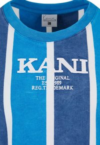 Mėlynas ir žydras dryžuotas megztinis su banguotu apykakle. Yra baltas siuvinėtas logotipas "KANI" ir tekstas "THE ORIGINAL EST. 1989 REG. TRADEMARK."