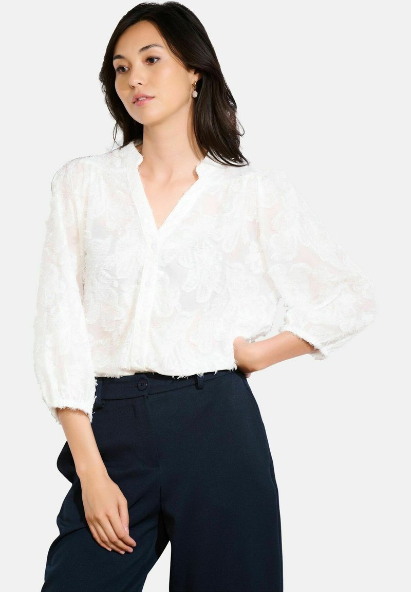 Blusa bianca con un motivo floreale testurizzato, scollatura a V e maniche a sbuffo. Abbinate a pantaloni scuri con chiusura a bottone.