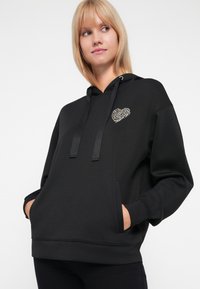 Fekete kapucnis pulóver, puha anyagból készült, bal mellen leopárdmintás szívvel, zsinóros kapucnival és elülső zsebekkel.