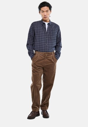 Homme portant une chemise à carreaux bleu marine boutonnée, un pantalon en velours côtelé marron et des chaussures marron foncé, debout les mains dans les poches.