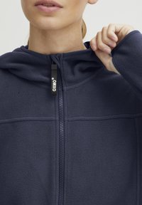 Sudadera con capucha de forro polar en color navy, con cierre de cremallera, que cuenta con una capucha ajustable y un tirador de cremallera con marca. Textura suave y costuras mínimas son visibles.