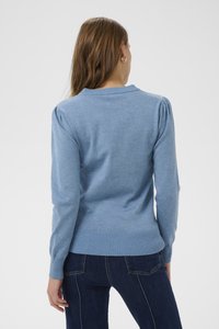 Nő hosszú barna hajjal, arany karika fülbevalókat visel, világoskék kötött pulóvert és sötétkék farmert, háttal fehér háttér előtt.