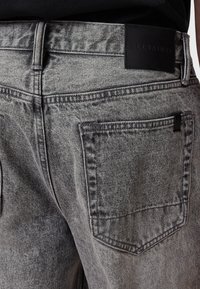 Gray denim jeans with a textured finish, featuring a black leather patch on the waistband and two back pockets with stitching details.