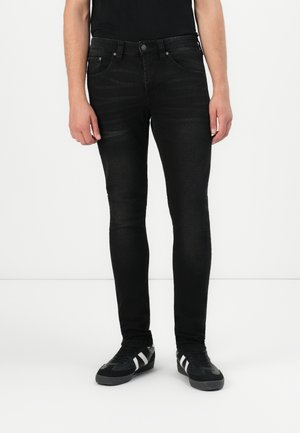 Jeans skinny negros con una textura suave, que presentan una cintura de altura media, bolsillos frontales y detalles de desvanecimiento sutil. Llevados con zapatos negros.