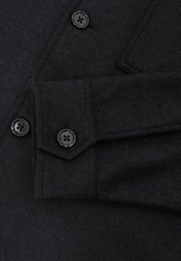 Fekete gyapjúpulóver testreszabott gallérral, két fekete gombbal és egy foltzsebbel az ujjon, texturált anyaggal.