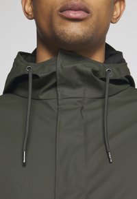 Homme portant une veste à capuche vert foncé avec des cordons et un col haut zippé jusqu'au cou.