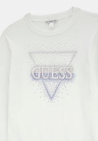 Pull blanc avec un col rond et un logo triangulaire orné de strass bleus et argentés, avec le mot « GUESS » affiché en évidence.