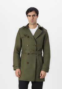 Trench coat verde oliva con frente de doble botonadura, botones marrones, detalle de cinturón y epaulettes. Hecho de una tela suave, diseño de longitud media.