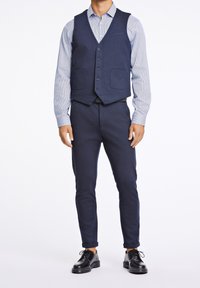 Marineblauer Weste über einem hellblau gestreiften Hemd, kombiniert mit navyblauen Hosen und schwarzen Schuhen. Die Weste hat einen Fünf-Knopf-Verschluss und zwei Vorderaschen.