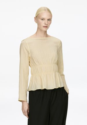 Hellgelbe Peplum-Bluse aus strukturiertem, kräuseligem Stoff, mit langen Ärmeln und gerafftem Bund, kombiniert mit schwarzen weiten Hosen.