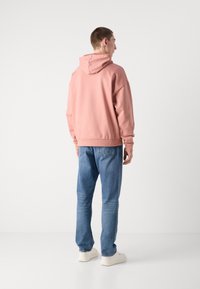 Rosa Kapuzenpullover mit Kordelzug, lässiger Schnitt und gerippten Bündchen, kombiniert mit blauen Jeans und weißen Sneakern. Rückansicht.