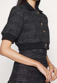 Svart tweed-kort jacka med korta ärmar, strukturerat tyg, ribbad nederkant, guldfärgade knappar och kontrasterande svart krage.