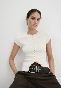 Femme aux cheveux foncés portant un haut blanc à manches courtes et boutonné ainsi qu'un pantalon foncé avec une large ceinture noire, debout devant un fond blanc uni.